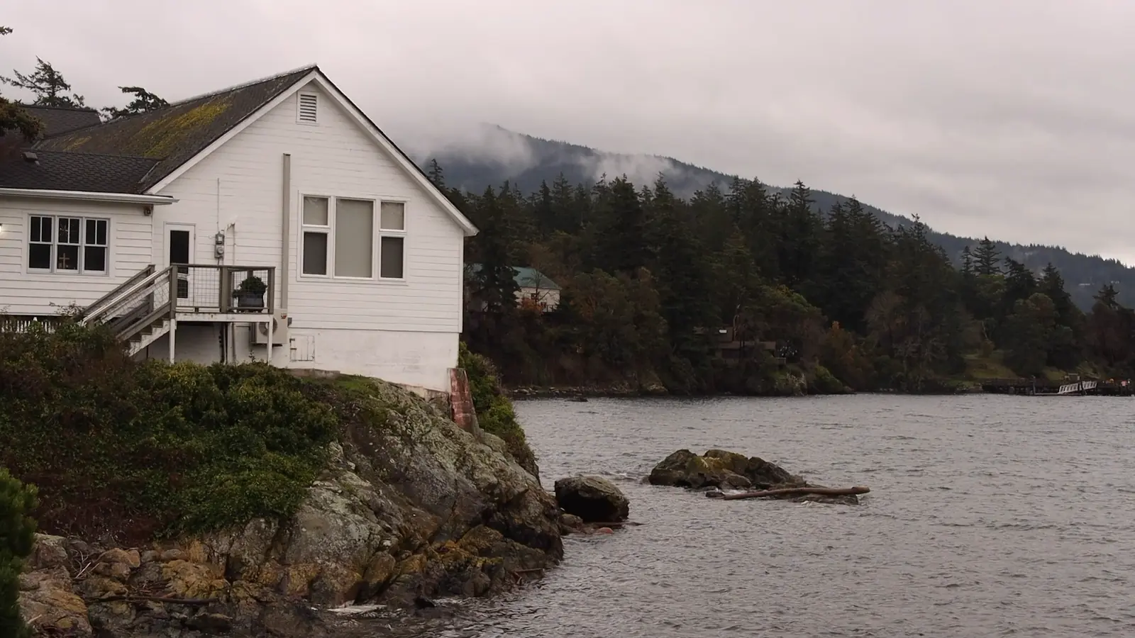 Still from documentary, white house on a rocky shore with fog-covered forested hills beyond